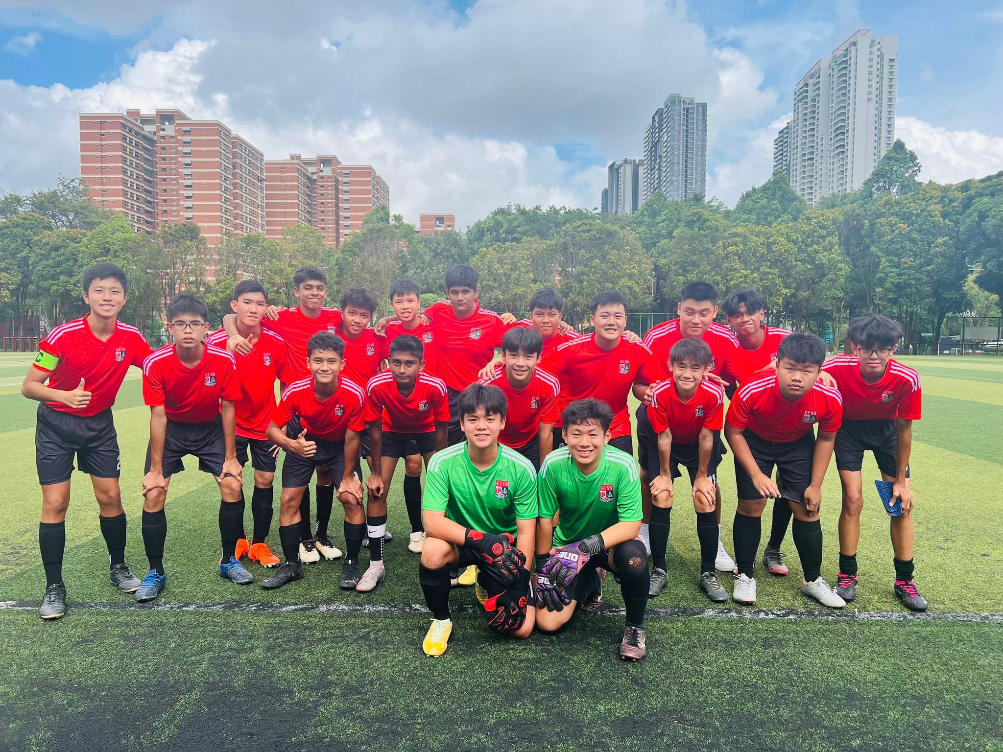 A youth soccer team in red jerseys poses on a green field, with two goalkeepers in green in front.